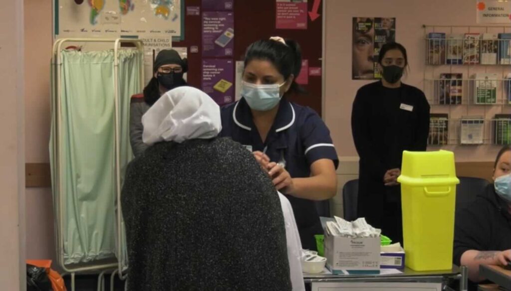 A nurse wearing a face mask giving an older lady a vaccination as several people wearing surgical face masks look on.