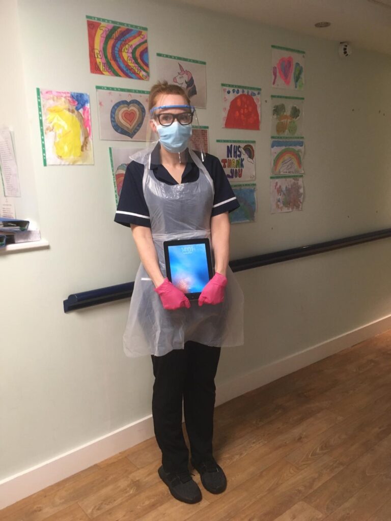 Lucy Walker in her nurse's uniform, wearing a face covering and face shield. She is holding an electronic tablet and gacing the camera.